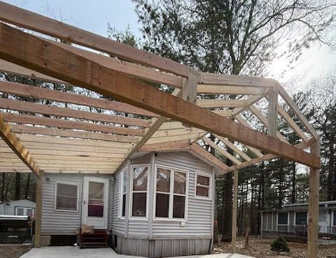 Under-construction porch framework attached to a mobile home surrounded by trees.