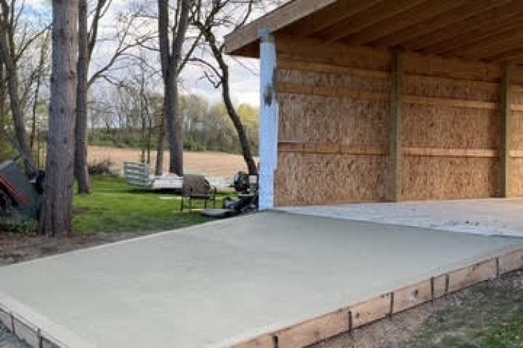 Freshly poured concrete slab at a wooden structure, surrounded by trees and outdoor seating.