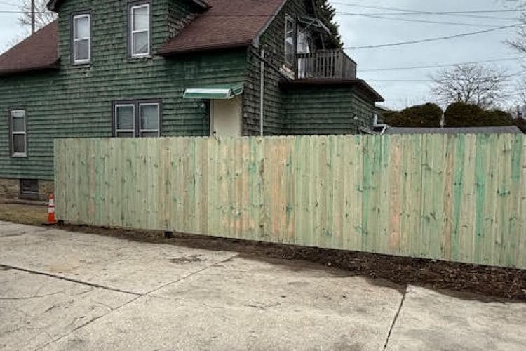 Green wooden fence in front of a house with a balcony and a paved driveway.