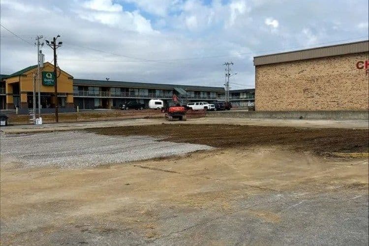 Vacant lot with gravel driveway near a motel and commercial building under cloudy sky.