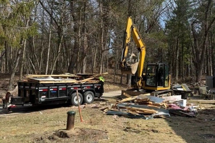 Excavator removing debris from property in wooded area, with trailer loaded with materials.