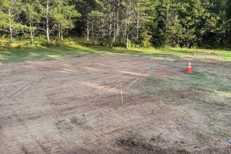 Cleared construction site showing dirt, survey marker, and safety cones in wooded area.