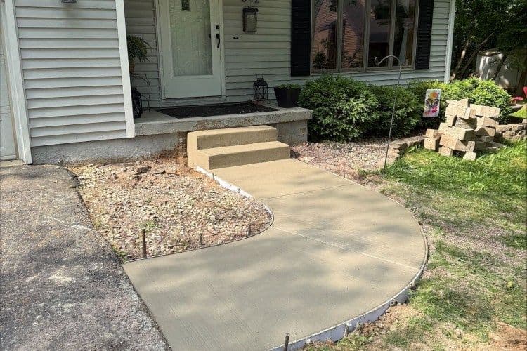 Newly poured concrete walkway with a step leading to a front door, landscaped surroundings.