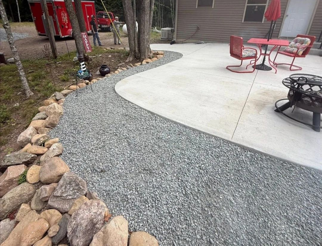 Gravel pathway leading to a patio area with a table and chairs, surrounded by rocks and trees.