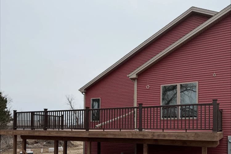 Red house with a spacious wooden deck and black railing under a cloudy sky.