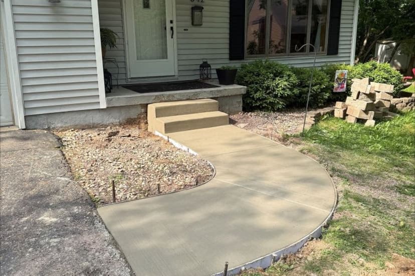 Newly poured concrete walkway leading to a front door, with landscaping elements nearby.