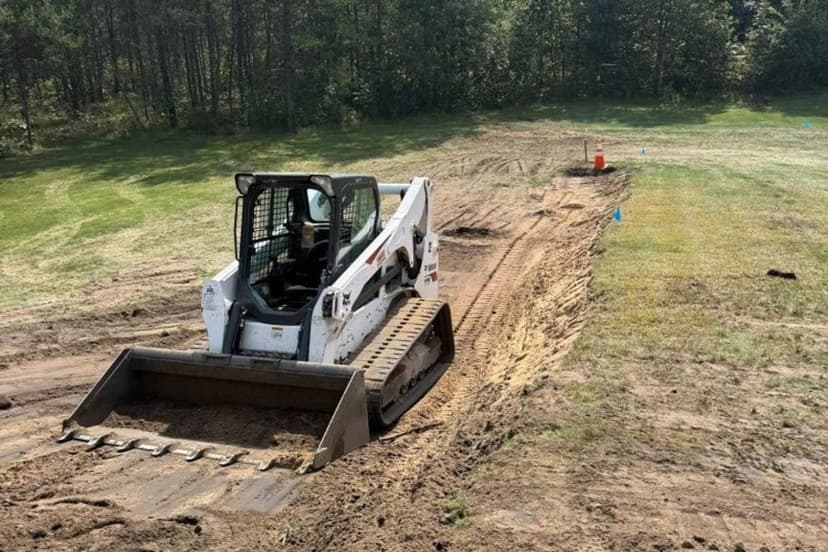 Bobcat skid steer leveling dirt on a construction site surrounded by trees.